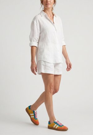 Vrouw draagt een wit overhemd met opgerolde mouwen, witte korte broek en kleurrijke sneakers met roze veters, lopend tegen een effen achtergrond.