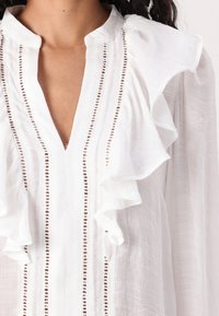 Weiße Bluse mit tiefem V-Ausschnitt, Rüschensäumen und dekorativen Details entlang der Vorderseite mit kleinen Öffnungen. Aus weichem, leichtem Stoff.