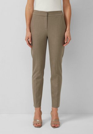 Femme portant un pantalon slim kaki longueur cheville à devant plat, associé à des sandales beiges à talons ouverts, sur un fond gris uni.