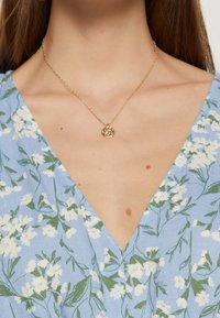 Collier en or avec un pendentif circulaire, présentant un design découpé. Porté par-dessus un tissu à motif floral bleu avec des accents blancs et verts.