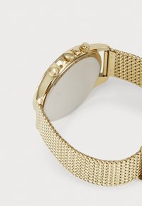 Montre en ton or avec un bracelet maillé, cadran rond et trois boutons couronne. Fini lisse et éclat métallique, avec un design minimaliste.
