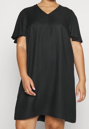 Kobieta w luźnej czarnej sukience z dekoltem w kształcie litery V, z krótkimi rozkloszowanymi rękawami i złotym pierścieniem na lewej ręce.