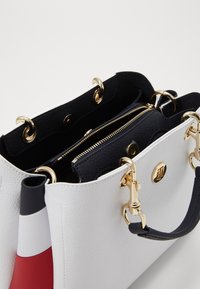 Hvid og sort taske med guld hardware, der har et lynlåslomme og farverige detaljer på den ene side. Glat overflade og struktureret form.