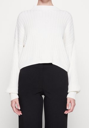 Pull en maille côtelée blanc à manches longues, associé à un pantalon noir taille haute, porté par une personne de la nuque jusqu'aux hanches.