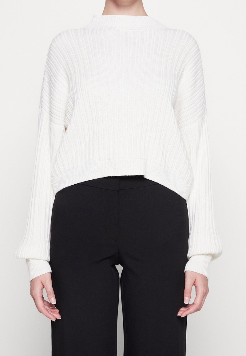Pull en maille côtelée blanc à manches longues, associé à un pantalon noir taille haute, porté par une personne de la nuque jusqu'aux hanches.