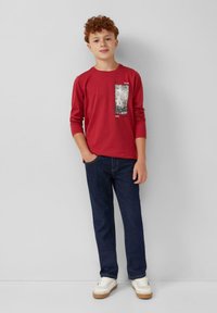 Rode lange mouw t-shirt met een grafische print op de voorkant, gecombineerd met donkere spijkerbroek. Witte sneakers met een rubberen zool maken de outfit compleet.