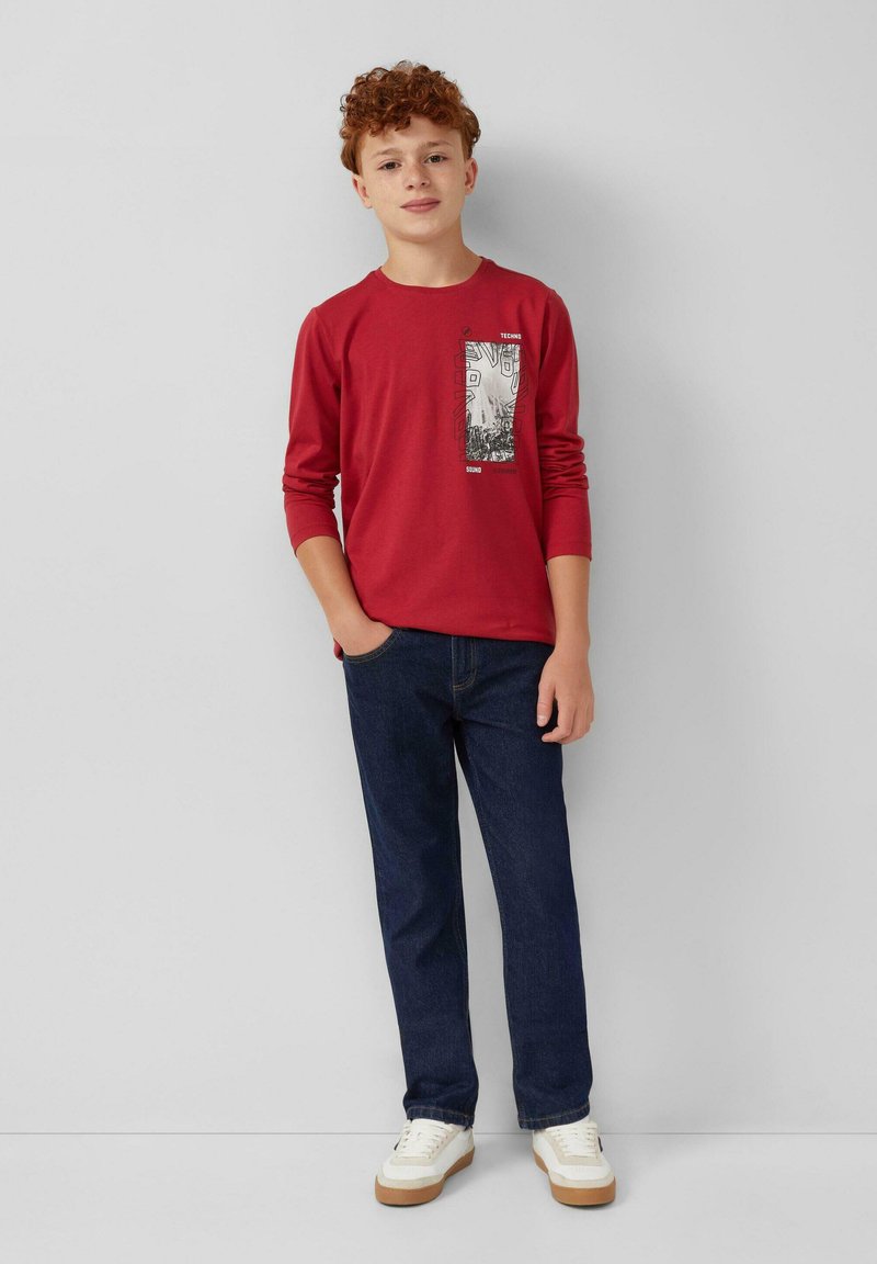 Rode lange mouw t-shirt met een grafische print op de voorkant, gecombineerd met donkere spijkerbroek. Witte sneakers met een rubberen zool maken de outfit compleet.