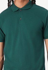 Dunkelgrünes Poloshirt mit klassischem Kragen, Knopfleiste mit drei Knöpfen und kurzen Ärmeln. Hergestellt aus strukturiertem Stoff und mit tailliertem Schnitt.