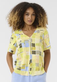 Īsām piedurknēm blūze ar V-veida kakla izgriezumu, ko rotā pasteļdzeltenā, zaļā un zilo ģeometrisko rakstu ar zeltainiem akcentiem un vaļīgu piegriezumu.