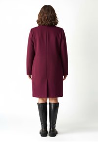 Donna con capelli castano ondulati che indossa un cappotto bordeaux fino al ginocchio e stivali neri alti fino al ginocchio, in piedi rivolta di schiena su uno sfondo bianco.