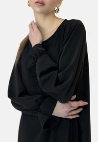 Schwarze Langarmbluse mit lockerer Passform, die geraffte Bündchen, eine glatte Textur und einen runden Halsausschnitt aufweist. Minimalistisches Design.
