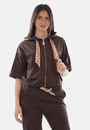 Donna che indossa una felpa marrone con zip, cordoncini beige con motivo e pantaloni marroni abbinati con cintura a cordoncino annodato.