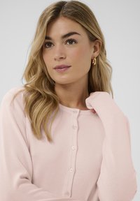 Svetlo roza rebrasti kardigan z okrolim ovratnikom, dolgimi rokavi in zapiranjem na sedem gumbov v enaki barvi. Gladka tekstura.