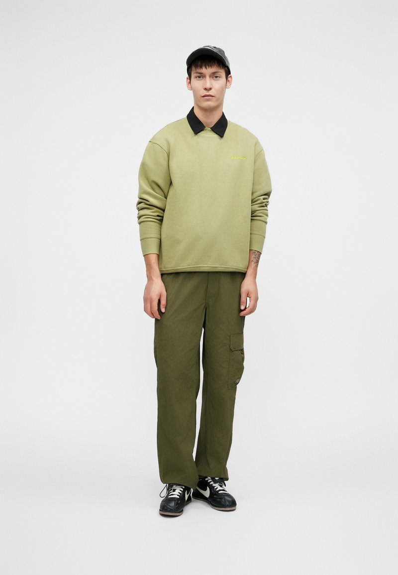 Sudadera verde con cuello negro, pantalones cargo en verde oscuro, zapatillas negras y una gorra gris. Presenta un logo bordado en el pecho.