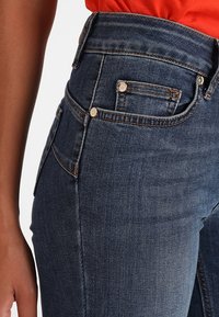 Jeans de denim azul oscuro de cintura alta con costuras sutiles, bolsillos frontales y botones de latón, que presentan una textura suave y una silueta ajustada.