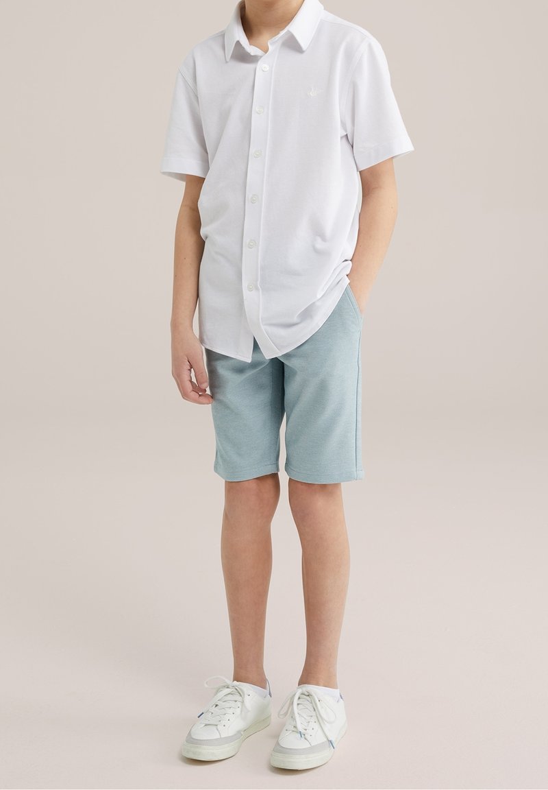 Garçon portant une chemise blanche à manches courtes boutonnée, un short bleu clair et des baskets blanches, debout avec une main dans la poche.