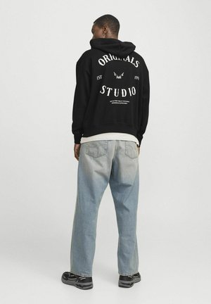 Black hoodie with white text, featuring "ORIGINALS" and "STUDIO" in a bold font. Worn with light blue, slightly distressed jeans.