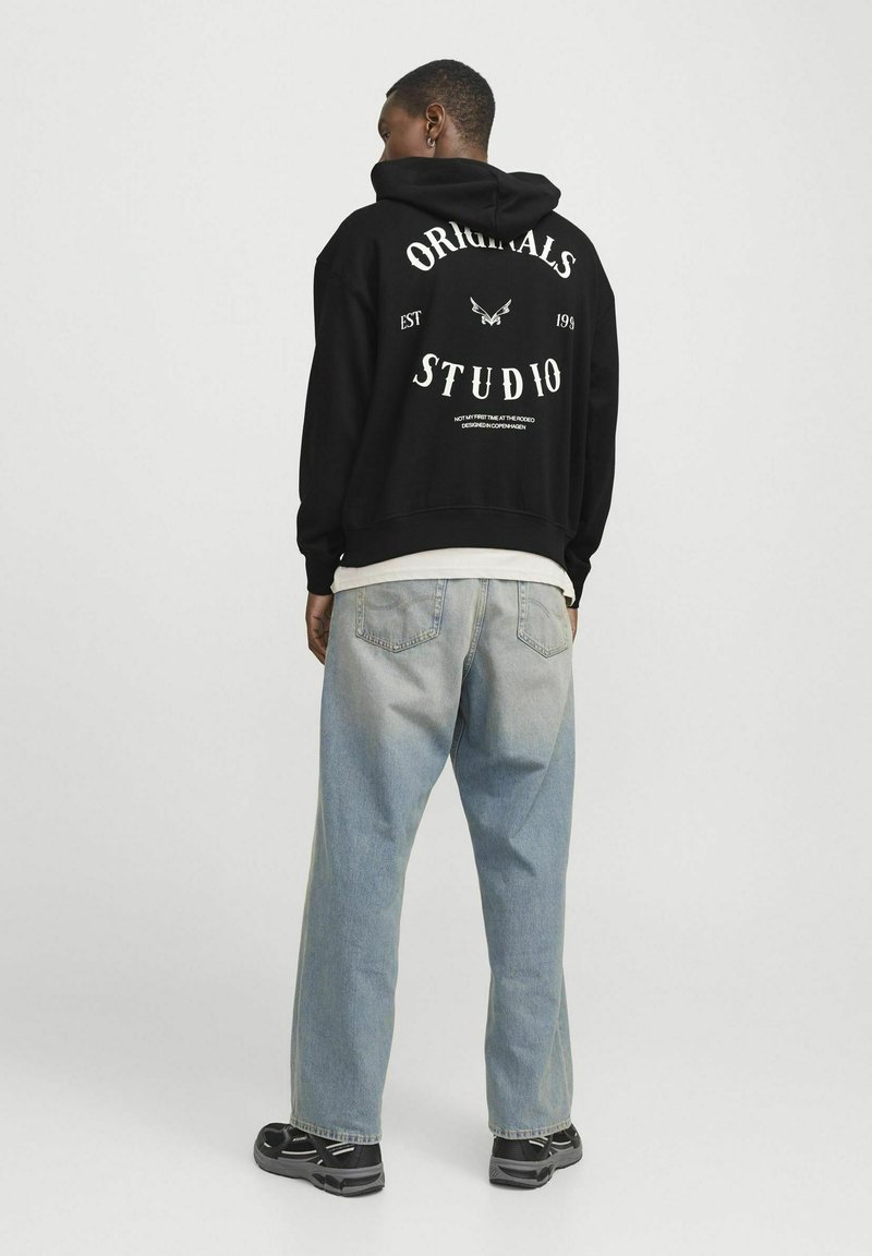 Zwarte hoodie met witte tekst, met de woorden "ORIGINALS" en "STUDIO" in een vet lettertype. Gekleed met lichtblauwe, iets versleten jeans.