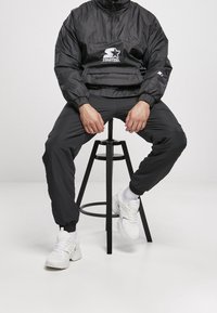 Black nylon windbreaker with a half-zip, white logo detail, paired with black joggers and white sneakers on a black stool.