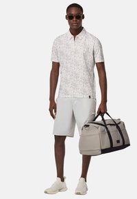 Chemise polo à motif floral léger, shorts beiges, sac de sport gris avec des accents noirs et chaussures de sport blanches.