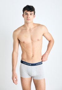 Γκρίζα μποξεράκια με ναυτική ζώνη που φέρει το λογότυπο "Lacoste". Ο ύφασμα φαίνεται λείος με εφαρμοστό σχέδιο. Το μοντέλο ποζάρει όρθιο.