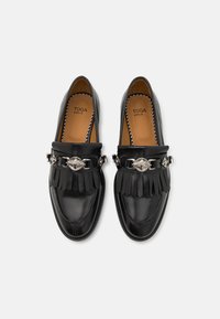 Mustad musta nahast loafers fringi detailide ja hõbedase dekoratiivse pandlaga, bränditud sisetallad tekstiga "TOGA VIRILIS," vaadet ülevalt.