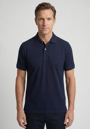 Mann mit hellbraunen Haaren, der ein marineblaues kurzärmeliges Poloshirt trägt, steht vor einem schlichten hellgrauen Hintergrund.
