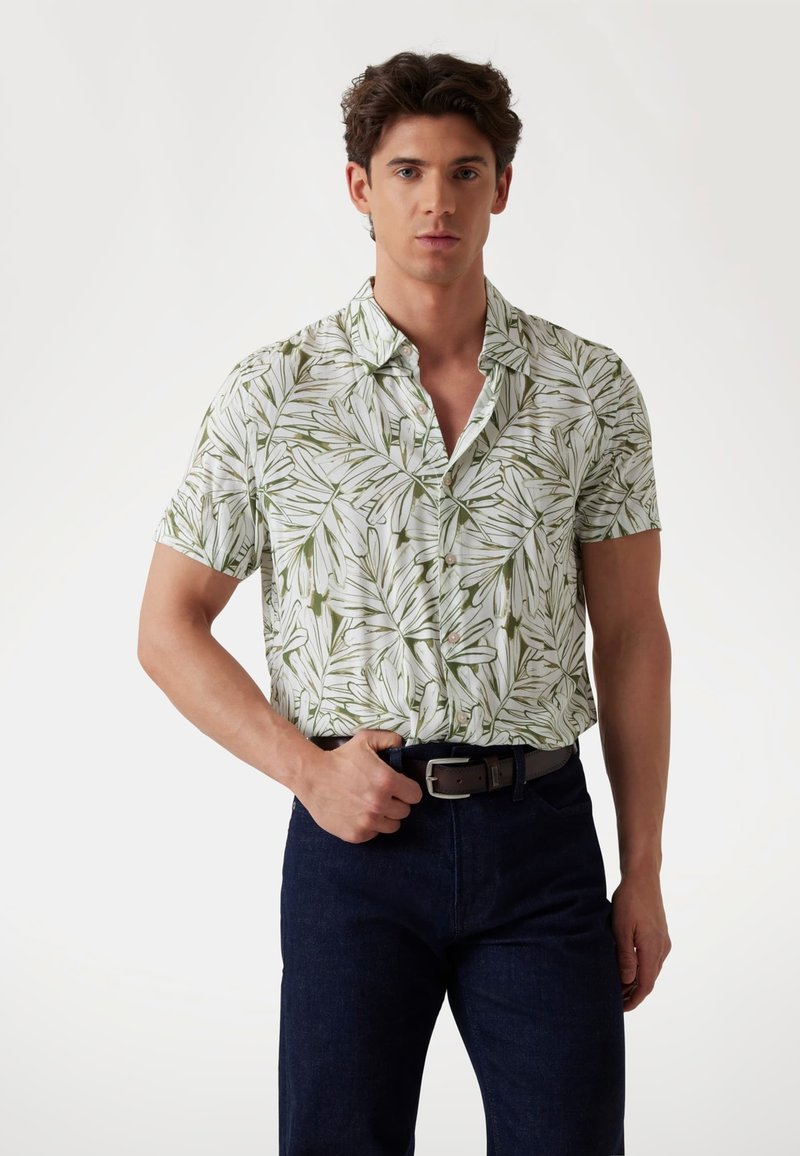 Camisa de manga corta con un patrón de hojas verdes sobre fondo blanco, confeccionada en tejido ligero, combinada con jeans oscuros y un cinturón marrón.
