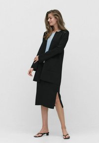 Svart kappa över en ljusblå topp, matchad med en svart kjol med en sidovan. Svarta sandaler fulländar outfiten. Släta texturer.
