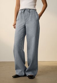 Jean large bleu clair avec une ceinture élastique et un cordon de serrage. Comprend des poches latérales, une texture lisse et une coupe décontractée.