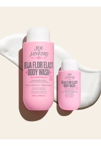 Deux flacons roses de Sol de Janeiro Beija Flor Elasti Body Wash, grands et format voyage, avec de la crème étalée en arrière-plan.