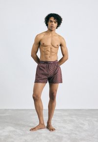 Calvin Klein rakstainie bokseršorti bordo krāsā, ar zīmola jostu, mīkstu audumu un brīvu piegriezumu, attēloti uz modeļa.