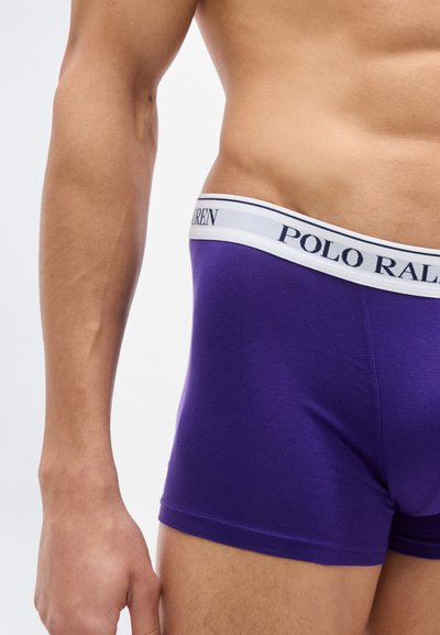 Αρσενικός κορμός και χέρι που φοράνε μοβ μπόξερ Polo Ralph Lauren με λευκή ζώνη μέσης με λογότυπο, σε λευκό φόντο.