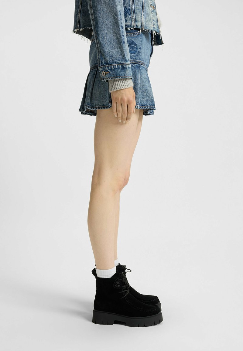 Persoană îmbrăcată cu o jachetă din denim și fustă, stând lateral, purtând șosete albe și cizme negre cu șireturi, pe un fundal simplu.