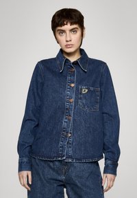 Ciemnoniebieska kurtka dżinsowa z rozłożystym kołnierzem, guzikami z przodu, kieszenią na piersi z naszywką z logo oraz długimi rękawami zakończonymi mankietami zapinanymi na guziki.