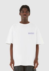 Hvid oversized t-shirt med korte ærmer, prydet med lilla tekstlogo på venstre brystområde. Glat bomuldsstof, afslappet pasform.