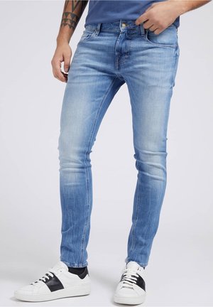 Homme portant un jean slim bleu clair et des baskets blanches avec des bandes noires, debout, une main près de la poche et un bras tatoué visible.