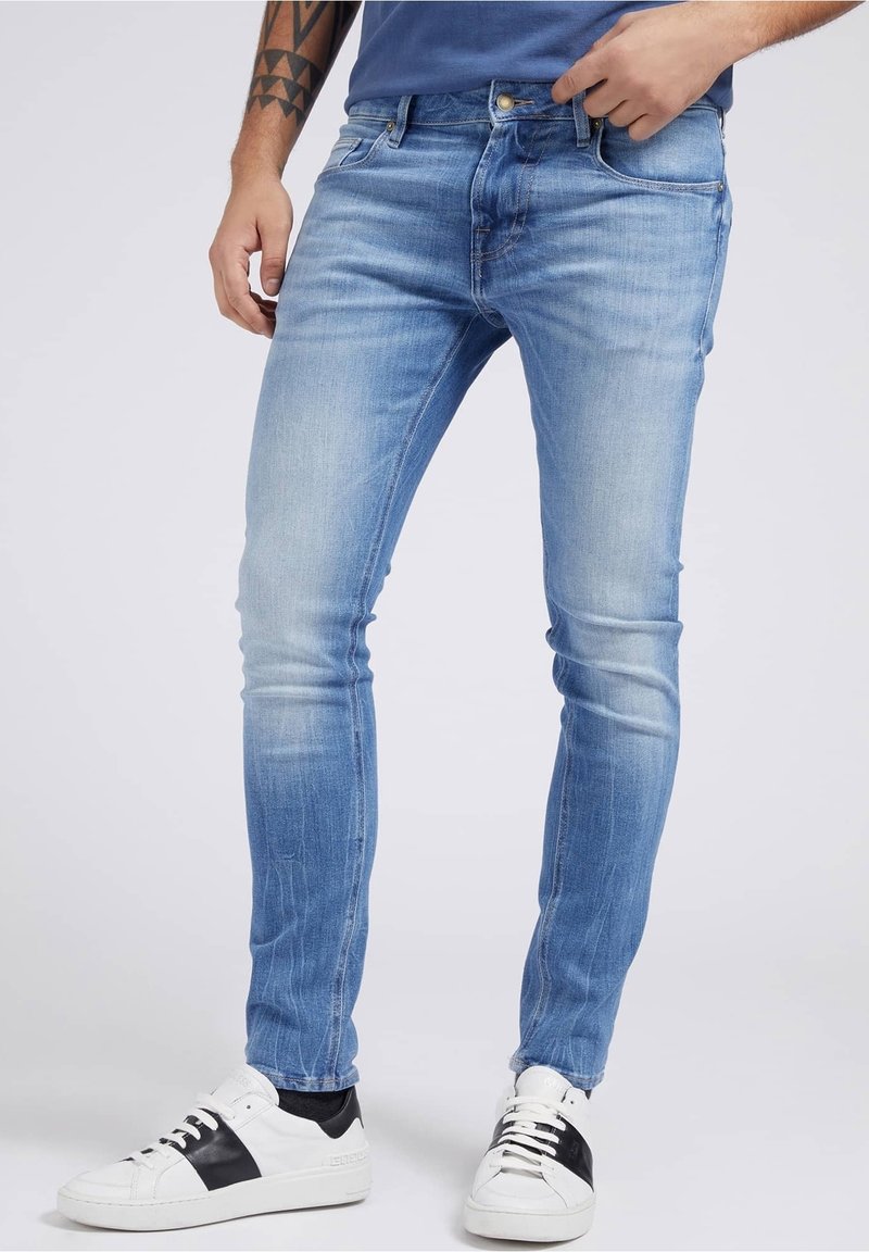 Homme portant un jean slim bleu clair et des baskets blanches avec des bandes noires, debout, une main près de la poche et un bras tatoué visible.