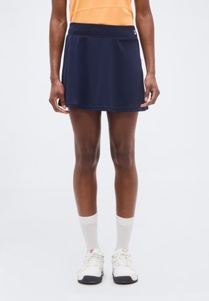 Persoon draagt een marineblauwe sportrok, witte sokken, witte sneakers en een oranje mouwloos shirt, staand tegen een effen achtergrond.