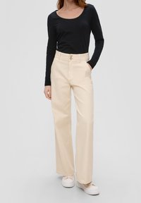 Beige weite Hose mit hohem Bund, ausgestattet mit einer doppelten Knopfverschluss und seitlichen Taschen. Kombiniert mit einem schwarzen Langarmoberteil und weißen Turnschuhen.