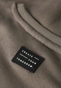 Zbliżenie brązowego materiału z żebrowanym szwem i czarną łatą z napisem "STWÓRZ SWOJE JUTRO" w białym tekście.