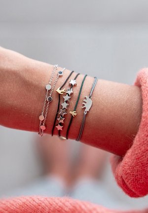 Ensemble de six bracelets sur un poignet : chaîne en argent, cordons colorés avec des motifs d'étoiles, d'oiseaux et d'un ours. Matériaux mélangés en argent, or et fil coloré.