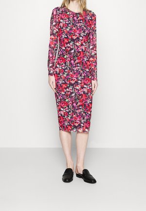Vrouw draagt een jurk met lange mouwen en knielengte met rood, roze en paarse bloemenprints, gecombineerd met zwarte instap loafers.