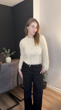 Crèmekleurige gebreide trui met ronde hals, knoopsluiting aan de voorkant en taille met trekkoord, gecombineerd met zwarte wijde broekspijpen.