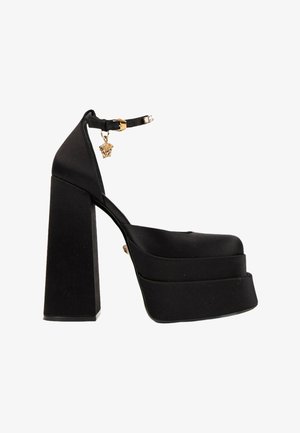 Chaussure à talon haut plateforme noire avec talon bloc épais, bride à la cheville et détail de pendentif doré près de la boucle.