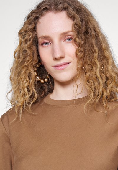 Brązowa koszulka w prążki z okrągłym dekoltem, wyróżniająca się delikatną teksturą. Duże, okrągłe, złote kolczyki w kształcie kół podkreślają stylizację, uwydatniając twarz.