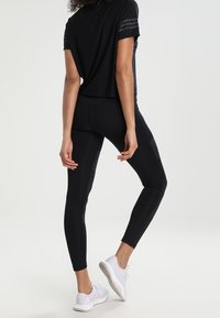 Maglietta sportiva nera a maniche corte con dettagli a righe, abbinata a leggings neri con una texture liscia. Scarpe sportive bianche completano l'outfit.