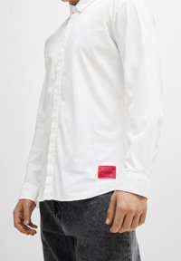 Witte lange mouwen shirt met knopenfront, voorzien van een klein rood logo-patch aan de linkerkant en een gladde katoenen textuur.