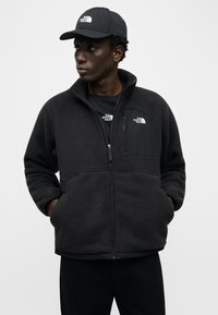 Νέος άνδρας που φοράει μαύρο φλις μπουφάν The North Face, αντίστοιχο καπέλο και μαύρο παντελόνι, στέκεται με τα χέρια στις τσέπες, κοιτάζοντας αριστερά.