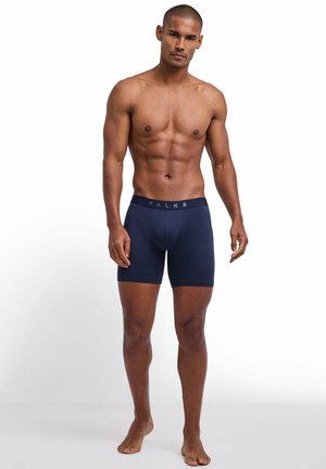 Męźczyzna o umięśnionej sylwetce stojący boso, ubrany w granatowe bokserki z paskiem z napisem „FALKE” na białym tle.